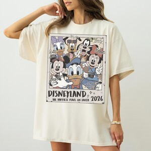 K&ouml;nnte beinhalten: Cremefarbenes T-Shirt mit einem Disneyland-Grafik im Vintage-Stil mit Micky Maus, Minnie Maus, Donald Duck und anderen Disney-Figuren. Die Grafik enth&auml;lt den Text "Disneyland" und "The Happiest Place on Earth 2026".