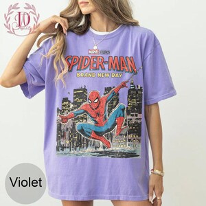 Puede incluir: Camiseta violeta con un gr&aacute;fico de Spider-Man de Marvel Studios. El dise&ntilde;o incluye el texto "SPIDER-MAN BRAND NEW DAY" y una ilustraci&oacute;n de Spider-Man contra un horizonte urbano. La camiseta tiene mangas cortas y cuello redondo.