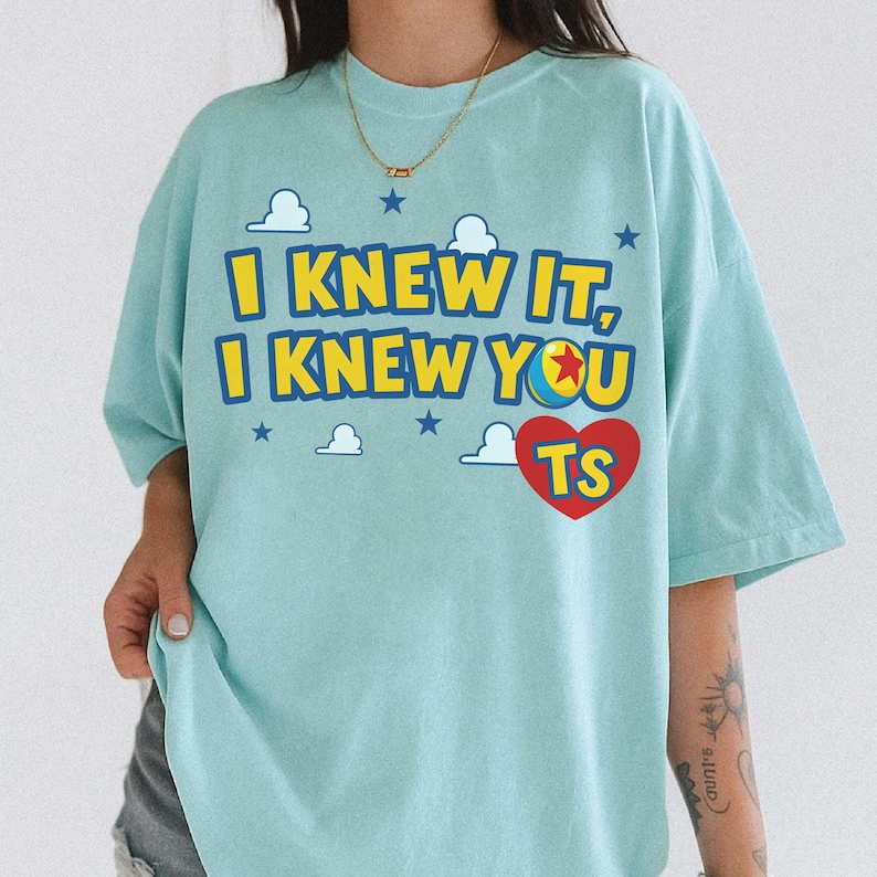 May include: A light blue t-shirt featuring the yellow text I KNEW IT, I KNEW YOU, a colourful ball graphic with a red star, and a red heart containing the letters TS. The design includes small white cloud and dark blue star icons throughout.