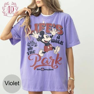 K&ouml;nnte beinhalten: Violettes T-Shirt mit Mickey Mouse und dem Text "LIFE'S A WALK IN THE PARK" und "Walt Disney World". Die Grafik enth&auml;lt eine Schlossillustration. Das Shirt hat kurze &Auml;rmel und einen Rundhalsausschnitt.
