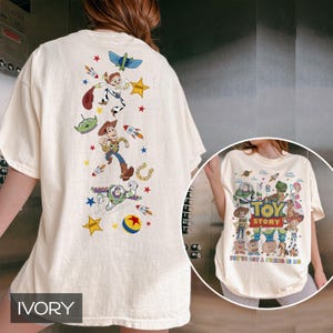 May include: Ivory-colored t-shirt featuring a colorful graphic of characters from the animated film "Toy Story." The front of the shirt displays the film's title and characters, while the back showcases additional characters and design elements.