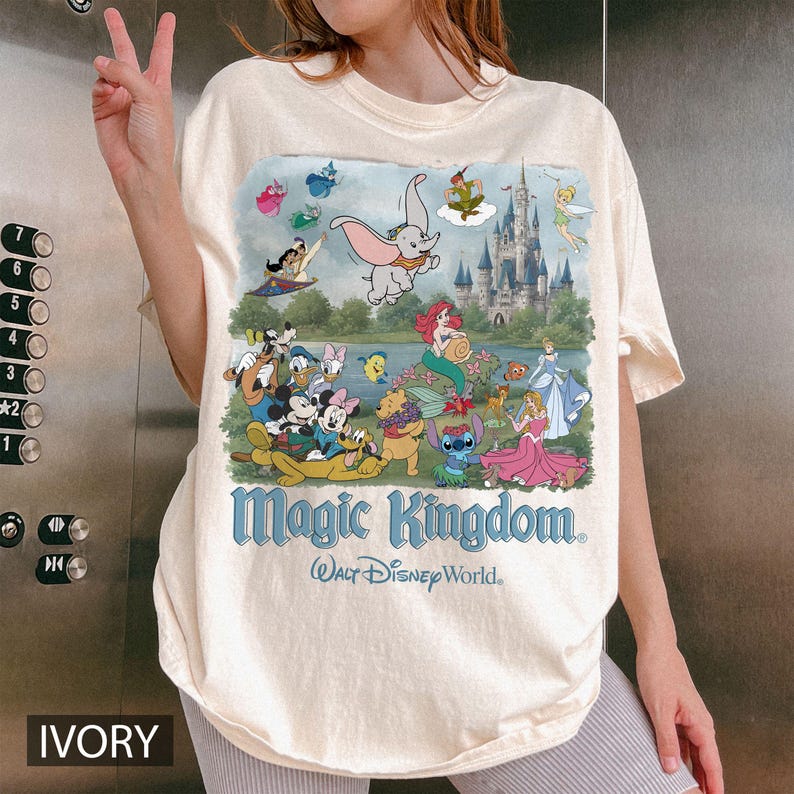May include: An ivory t-shirt featuring a colorful graphic print of various Disney characters including Dumbo, Mickey Mouse, and Ariel in front of a castle. The text Magic Kingdom and Walt Disney World is printed in blue below the character illustration.