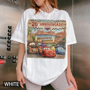 Puede incluir: Una camiseta blanca con un estampado gr&aacute;fico que celebra el 20&ordm; aniversario de la pel&iacute;cula Cars. El dise&ntilde;o muestra personajes de Radiator Springs, incluidos Rayo McQueen y Mate, sobre un fondo de paisaje des&eacute;rtico.