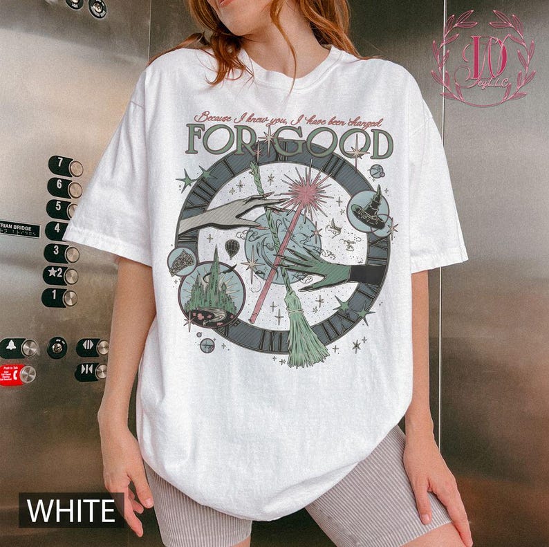 Puede incluir: Camiseta blanca con un gr&aacute;fico caprichoso que presenta un dise&ntilde;o de reloj, manos, un castillo y un sombrero de bruja. El texto "FOR GOOD" se muestra de forma destacada, con la frase "Because I know you, I have been changed" encima. La camiseta tiene mangas cortas.