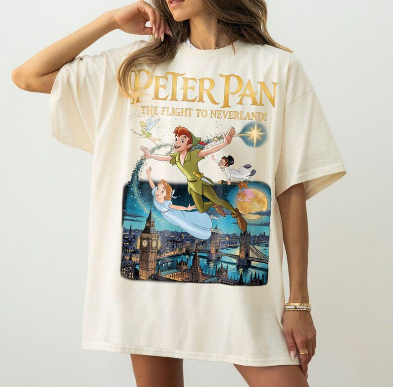 May include: Cream-coloured t-shirt with a colourful graphic of Peter Pan, Wendy, and Tinkerbell flying over London. The text "PETER PAN" and "THE FLIGHT TO NEVERLAND!" are printed in gold. The shirt has short sleeves.