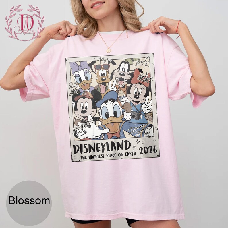 K&ouml;nnte beinhalten: Hellrosa T-Shirt mit einem Retro-Motiv von Disney-Figuren, darunter Micky Maus und Donald Duck. Der Aufdruck lautet "Disneyland" und "The Happiest Place on Earth 2026". Das T-Shirt hat eine lockere Passform.