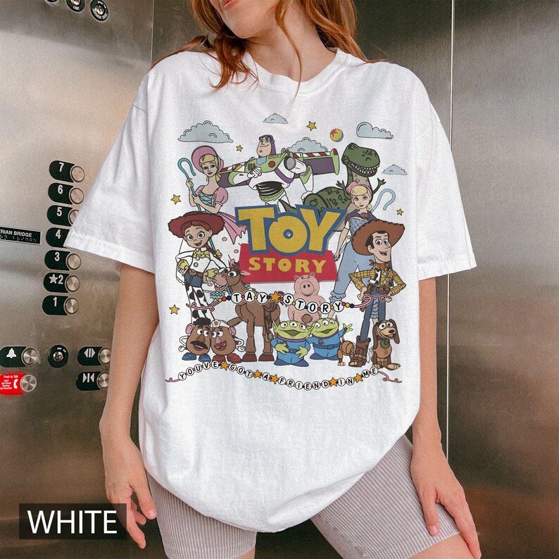 May include: A white short-sleeve t-shirt featuring a colorful graphic print of Toy Story characters including Woody, Buzz Lightyear, Jessie, and others. The design includes the text "TOY STORY" in large letters and "YOU'VE GOT A FRIEND IN ME" below.
