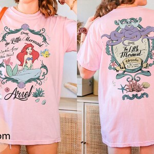 May include: A light pink t-shirt featuring a graphic of Ariel from Disney's The Little Mermaid. The front design includes Ariel, Flounder, and the text "The Little Mermaid". The back features Ursula. The shirt is a comfortable, casual style.