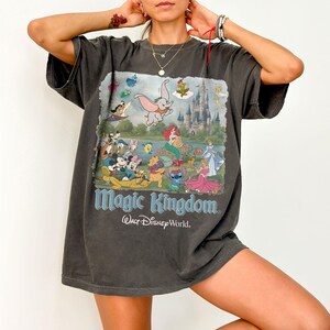 May include: A charcoal gray short-sleeve t-shirt featuring a large graphic print of various Disney characters, including Dumbo, Mickey Mouse, and Ariel, in front of a castle. The text Magic Kingdom and Walt Disney World is printed below the graphic.