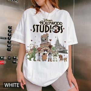 Pode incluir: Camiseta branca com o texto "Disney's Hollywood Studios" e um gr&aacute;fico de personagens e atra&ccedil;&otilde;es do Toy Story. A palavra "WHITE" est&aacute; impressa na parte inferior da camiseta.
