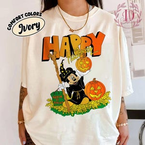Peut inclure: T-shirt ivoire avec un motif d'Halloween. Le dessin représente Mickey Mouse déguisé en sorcier tenant une citrouille et un bâton, avec les mots "HAPPY Halloween" en orange et noir.