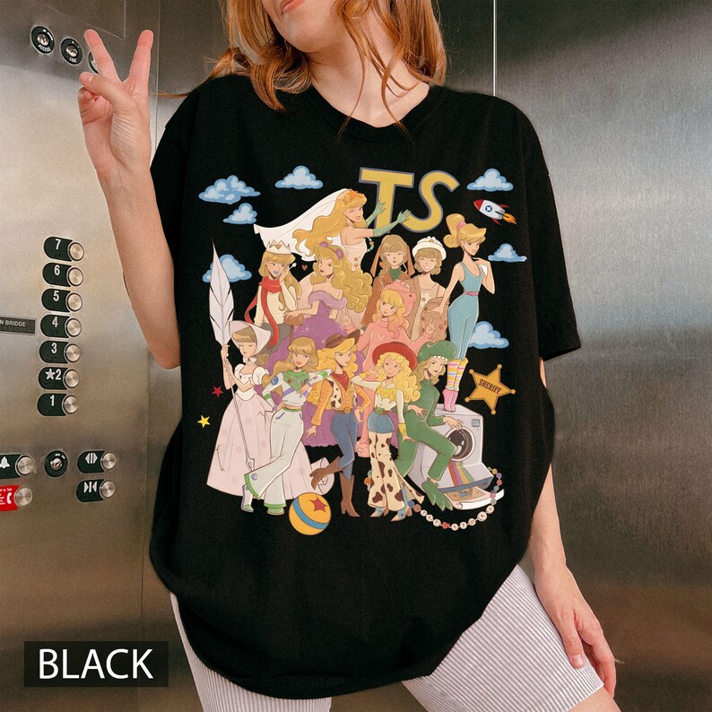 May include: A black t-shirt featuring a graphic print of multiple blonde characters in various outfits, including a space suit, cowgirl attire, and formal gowns. The design includes the letters TS, blue clouds, a rocket, and a sheriff star.