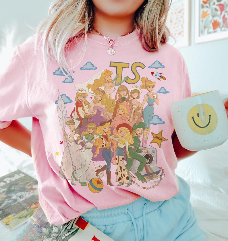 May include: A light pink t-shirt featuring a graphic print of various animated characters in diverse outfits, including a space ranger suit and cowboy attire. The design includes the letters TS, a rocket, clouds, a star-shaped badge, and a camera.