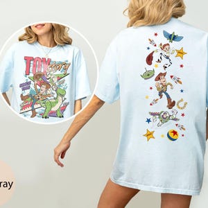 May include: Pale blue t-shirt with a Toy Story graphic. The front has the film's title and characters, the back features a vertical design of characters and rockets. A relaxed, casual top.