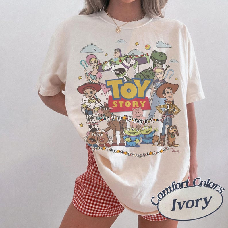 May include: An ivory short-sleeve t-shirt featuring a colorful graphic print of Toy Story characters including Woody, Buzz Lightyear, Jessie, and others. The design includes the Toy Story logo and the text I've Got A Friend In Me in a circular pattern.