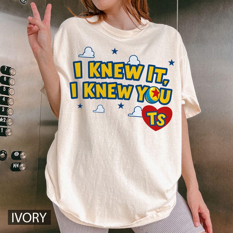 May include: An ivory short-sleeve t-shirt featuring the blue and yellow text I KNEW IT, I KNEW YOU with a colourful ball graphic, a red heart containing the letters TS, and small blue cloud and star icons printed on the front chest area.
