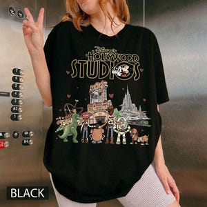 Pode incluir: Camiseta preta com o texto "Disney's Hollywood Studios" e personagens de desenhos animados. O design inclui Mickey Mouse e imagens de atra&ccedil;&otilde;es. A palavra "BLACK" est&aacute; impressa na parte inferior.