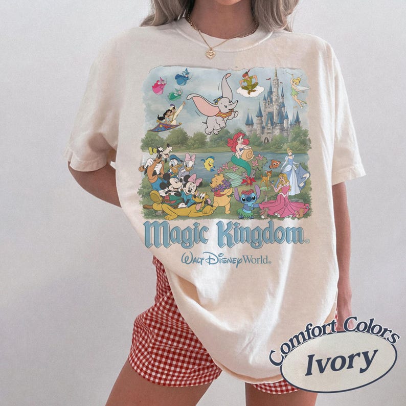 May include: An ivory short-sleeve t-shirt featuring a colorful graphic print of various animated characters, a castle, and the text Magic Kingdom Walt Disney World. The shirt is paired with red and white checkered shorts and a gold chain necklace.