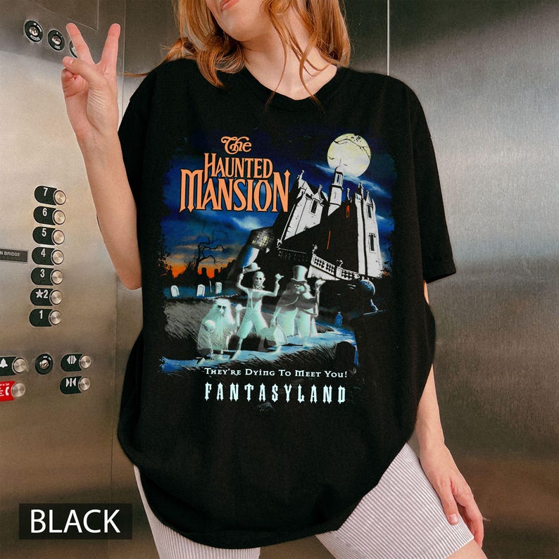 May include: Black t-shirt featuring a graphic from The Haunted Mansion ride at Disneyland. The design includes the ride's logo, a spooky mansion, and ghostly figures. The text "They're Dying To Meet You!" and "FANTASYLAND" are also visible.