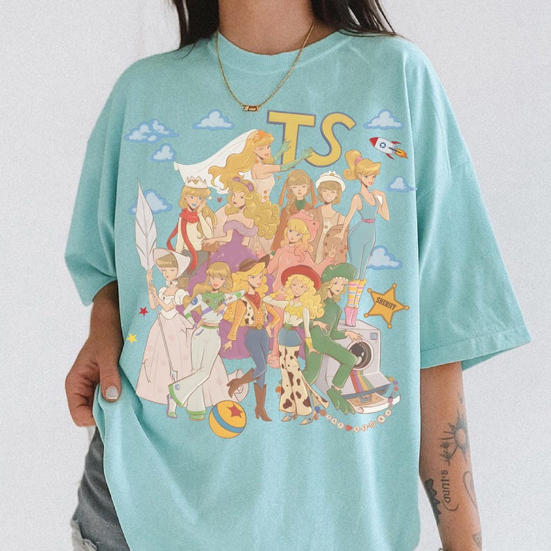 May include: A light blue t-shirt featuring a graphic print of various animated characters in a collage style. The design includes the letters TS, a rocket, a sheriff star, a ball, and a camera. The characters are depicted with blonde hair.