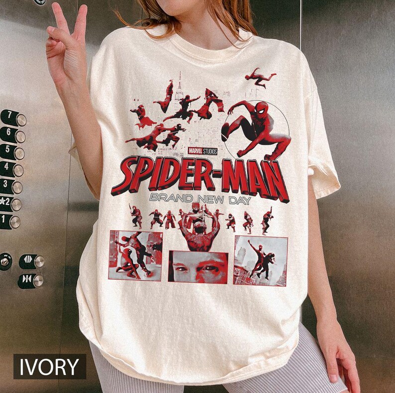 May include: Ivory-colored t-shirt featuring the Spider-Man logo and graphics from the movie. The design includes the text "SPIDER-MAN BRAND NEW DAY" in red. The shirt has a crew neck and short sleeves.