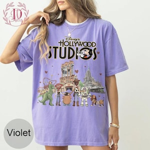 Pode incluir: Camiseta violeta com o texto "Disney's Hollywood Studios" e um gr&aacute;fico do Mickey Mouse e personagens de parques tem&aacute;ticos. O design inclui ilustra&ccedil;&otilde;es de atra&ccedil;&otilde;es. A camiseta tem gola redonda e mangas curtas.