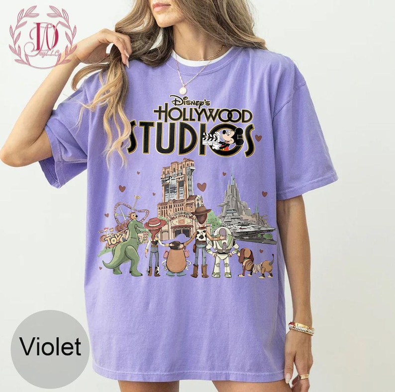 Pode incluir: Camiseta violeta com o texto "Disney's Hollywood Studios" e um gr&aacute;fico do Mickey Mouse e personagens de parques tem&aacute;ticos. O design inclui ilustra&ccedil;&otilde;es de atra&ccedil;&otilde;es. A camiseta tem gola redonda e mangas curtas.