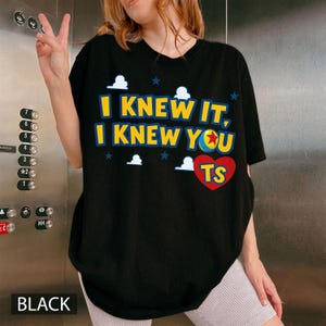 May include: A black short-sleeved t-shirt featuring the yellow text I KNEW IT, I KNEW YOU, a colourful ball graphic, and a red heart with the letters TS. The design includes small white cloud shapes and blue stars scattered across the front.