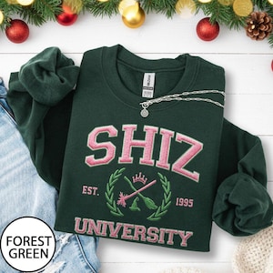 May include: Forest green sweatshirt with pink embroidered text reading "SHIZ UNIVERSITY". The design features a crest with crossed brooms and the text "EST. 1995". A silver necklace is draped across the sweatshirt.