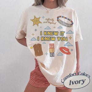 Puede incluir: Una camiseta color marfil con un estampado gr&aacute;fico que incluye el texto I KNEW IT I KNEW YOU, infinity and beyond, y YOU'VE GOT A FRIEND IN ME. El dise&ntilde;o presenta una estrella de sheriff amarilla, una pulsera de cuentas, botas y un sombrero rojo.