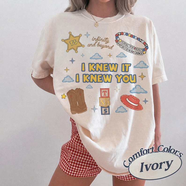 Puede incluir: Una camiseta color marfil con un estampado gr&aacute;fico que incluye el texto I KNEW IT I KNEW YOU, infinity and beyond, y YOU'VE GOT A FRIEND IN ME. El dise&ntilde;o presenta una estrella de sheriff amarilla, una pulsera de cuentas, botas y un sombrero rojo.
