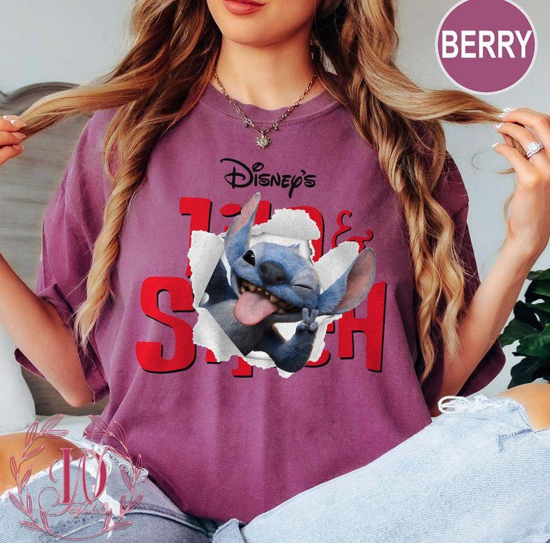 Op de afbeelding: Bes-kleurig T-shirt met het Disney-personage Stitch dat door het ontwerp van het shirt breekt. Het shirt heeft de tekst "Disney's" boven het personage en "Stitch" in rode blokletters.
