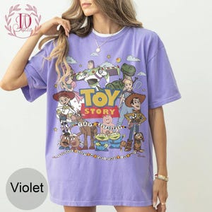 May include: A violet short-sleeved t-shirt featuring a graphic print of Toy Story characters including Woody, Buzz Lightyear, Jessie, Rex, and others. The text Toy Story and You've Got A Friend In Me are printed on the front with colorful star accents.