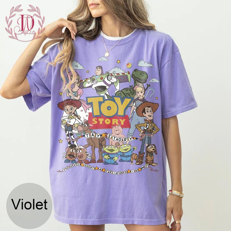 May include: A violet short-sleeved t-shirt featuring a graphic print of Toy Story characters including Woody, Buzz Lightyear, Jessie, Rex, and others. The text Toy Story and You've Got A Friend In Me are printed on the front with colorful star accents.