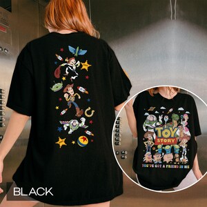 May include: Black t-shirt featuring colorful graphics of characters from the animated film "Toy Story." The back of the shirt displays Woody, Buzz Lightyear, and other characters with rockets and stars. The front of the shirt has the words "Toy Story" and "You've Got a Friend in Me."