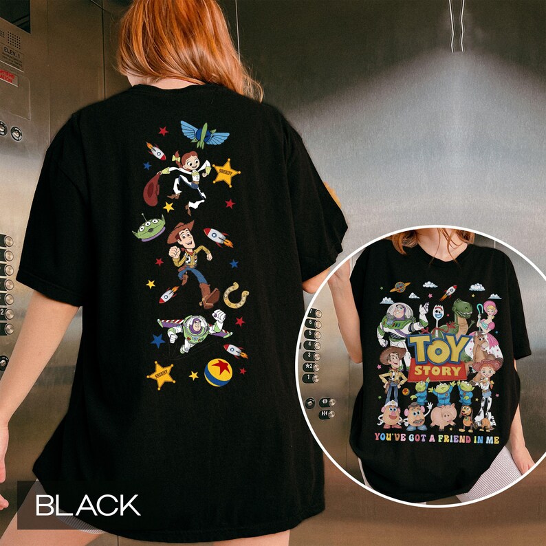 May include: Black t-shirt featuring colorful graphics of characters from the animated film "Toy Story." The back of the shirt displays Woody, Buzz Lightyear, and other characters with rockets and stars. The front of the shirt has the words "Toy Story" and "You've Got a Friend in Me."