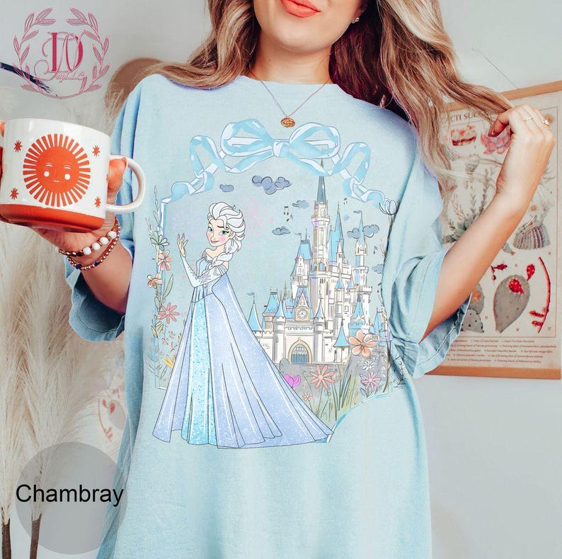 May include: Light blue t-shirt with a graphic of a princess in a blue dress and a castle. The shirt has a large light blue bow and the word "Chambray" at the bottom.