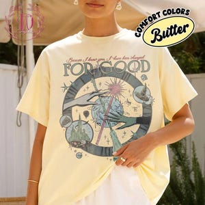 Puede incluir: Camiseta amarilla mantequilla con un dise&ntilde;o gr&aacute;fico que presenta un reloj, manos y una varita m&aacute;gica. El texto "FOR GOOD" se muestra de forma destacada, con texto adicional en la parte superior. La camiseta tiene mangas cortas y un corte holgado.