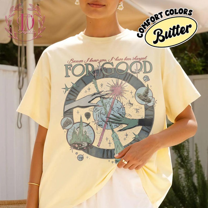 Puede incluir: Camiseta amarilla mantequilla con un dise&ntilde;o gr&aacute;fico que presenta un reloj, manos y una varita m&aacute;gica. El texto "FOR GOOD" se muestra de forma destacada, con texto adicional en la parte superior. La camiseta tiene mangas cortas y un corte holgado.