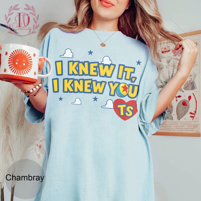 May include: A light blue t-shirt featuring the yellow text I KNEW IT, I KNEW YOU with a colourful ball graphic and a red heart containing the letters TS. The design includes white clouds and blue stars. A white mug with a red sun graphic is held nearby.