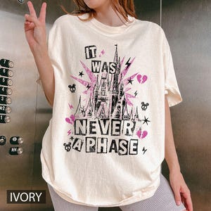 May include: Ivory-colored t-shirt with a graphic design featuring a castle, lightning bolts, stars, and the text "IT WAS NEVER A PHASE" in black and pink. The shirt has short sleeves and a relaxed fit.