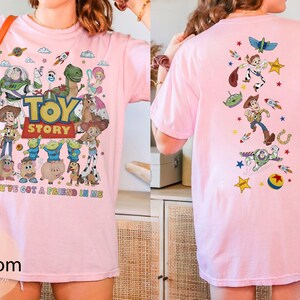 May include: Pink t-shirt featuring the Toy Story logo and characters like Woody, Buzz Lightyear, and Rex. The front of the shirt displays the phrase "You've Got a Friend in Me." The back of the shirt has a design with various Toy Story characters and elements.