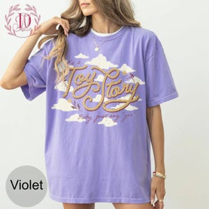 May include: A violet short-sleeve t-shirt featuring a graphic of white clouds and the text "It's a Toy Story" written in a rope-style font, with the additional script text "Baby, just say yes" printed below the main graphic design.