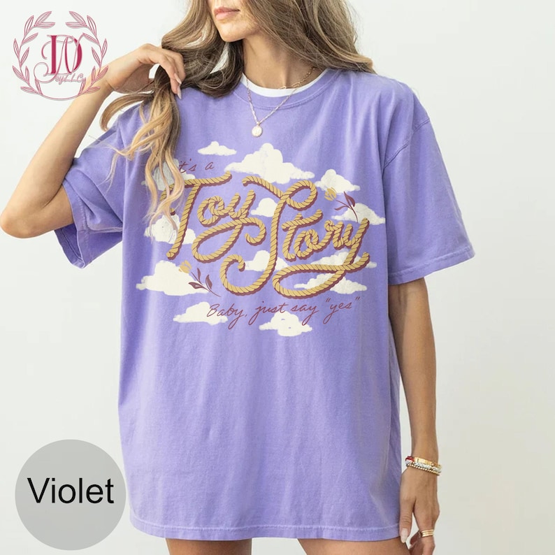 May include: A violet short-sleeve t-shirt featuring a graphic of white clouds and the text "It's a Toy Story" written in a rope-style font, with the additional script text "Baby, just say yes" printed below the main graphic design.