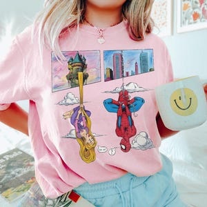 Peut inclure: Sweat-shirt rose clair avec un motif de bande dessin&eacute;e repr&eacute;sentant Raiponce et Spider-Man. Raiponce est suspendue &agrave; ses cheveux et Spider-Man est &agrave; l'envers. Une tasse blanche avec un visage souriant jaune est tenue.