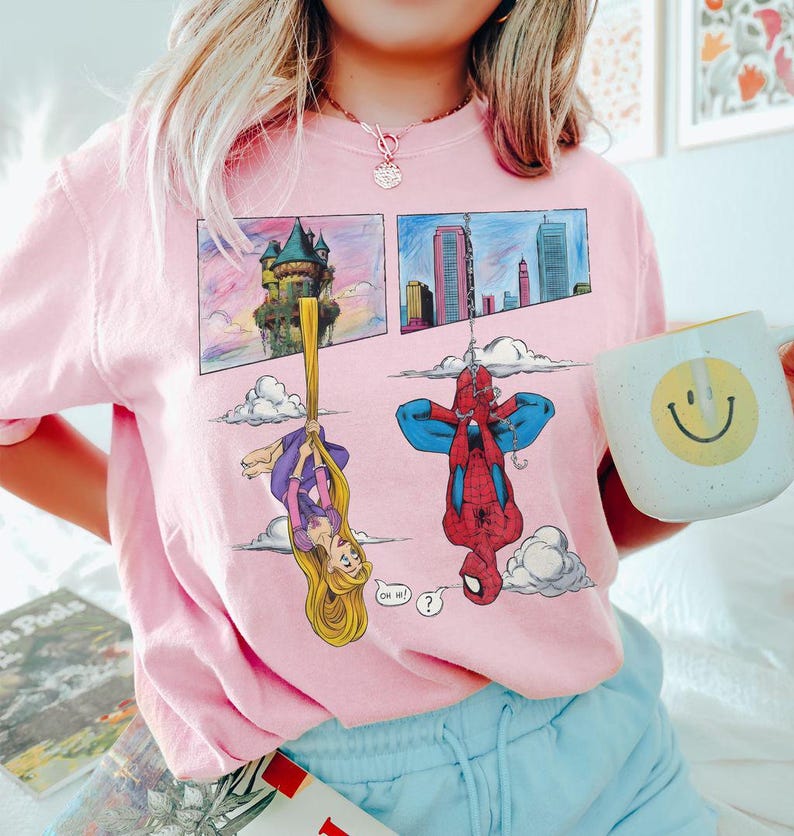 Peut inclure: Sweat-shirt rose clair avec un motif de bande dessin&eacute;e repr&eacute;sentant Raiponce et Spider-Man. Raiponce est suspendue &agrave; ses cheveux et Spider-Man est &agrave; l'envers. Une tasse blanche avec un visage souriant jaune est tenue.