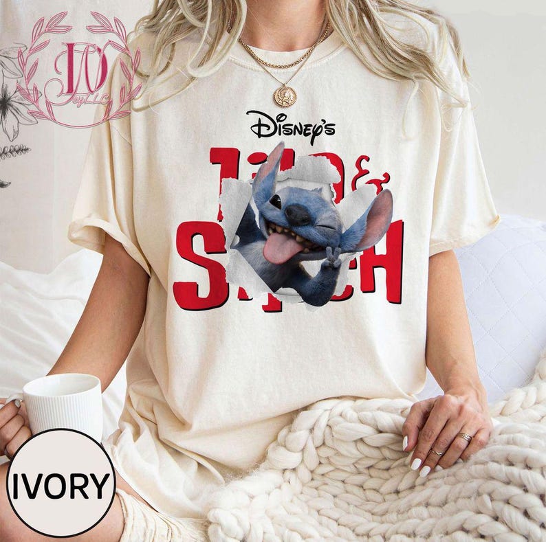 Op de afbeelding: Cr&egrave;mekleurig T-shirt met de tekst "Disney's" boven "100 & Stitch" in het rood. Een blauw Stitch-personage breekt door de afbeelding van het shirt. Het T-shirt heeft korte mouwen.