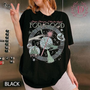 Puede incluir: Camiseta negra con un dise&ntilde;o gr&aacute;fico que presenta un reloj, manos, una varita m&aacute;gica y el texto "FOR GOOD". El dise&ntilde;o incluye estrellas, un castillo y un sombrero de bruja. El texto "Because I knew you, I have been changed" est&aacute; arriba.