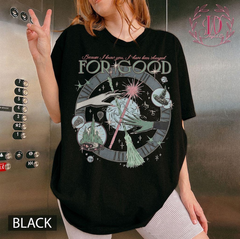 Puede incluir: Camiseta negra con un dise&ntilde;o gr&aacute;fico que presenta un reloj, manos, una varita m&aacute;gica y el texto "FOR GOOD". El dise&ntilde;o incluye estrellas, un castillo y un sombrero de bruja. El texto "Because I knew you, I have been changed" est&aacute; arriba.