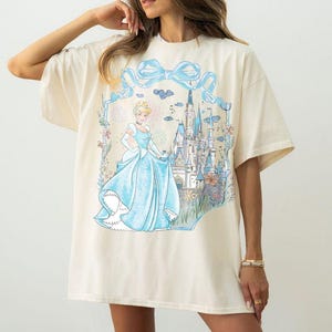 May include: Cream-colored t-shirt featuring a Cinderella graphic. The design includes Cinderella in a blue gown, a castle, and a blue bow. The shirt has short sleeves and a relaxed fit.
